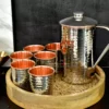 Hammered stainless steel and copper pitcher and tumbler set on a carved wooden tray for a premium kitchenware look.