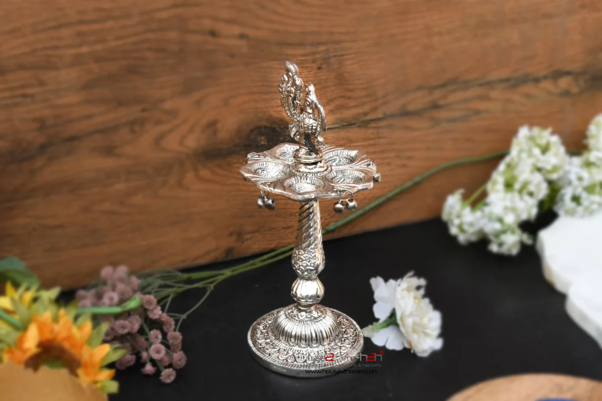 Ornate German silver peacock diya stand with hanging metal bells placed against a wooden backdrop.