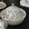 decorative silver metal bowl with an elaborate openwork design, resting on a white lotus-shaped base.