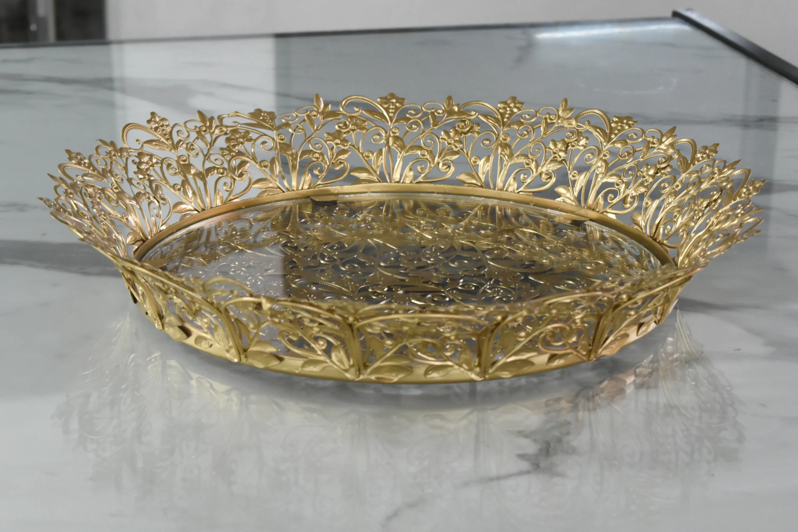 Gold-finished filigree metal round tray with intricate lace border and glass base for home styling.