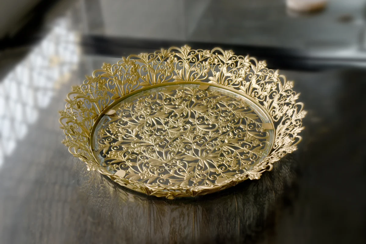 Gold-finished filigree metal round tray with intricate lace border and glass base for home styling.