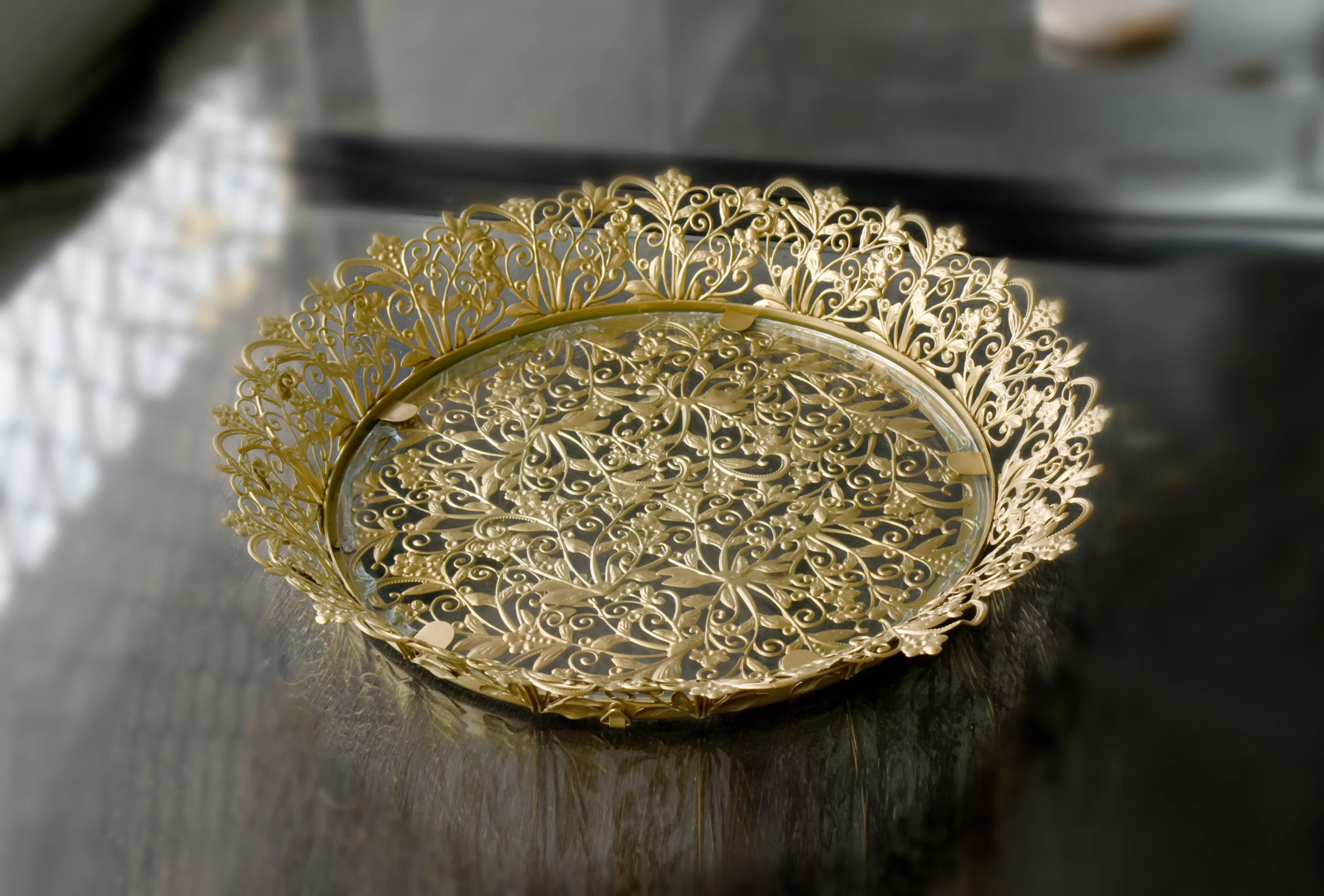 Gold-finished filigree metal round tray with intricate lace border and glass base for home styling.